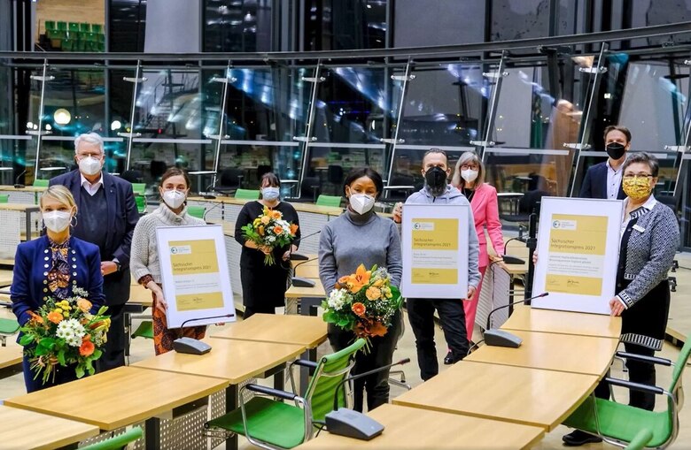 Gruppe von neun Personen in einem modernen Sitzungssaal, einige mit Mund-Nasen-Schutz, halten Blumen und Urkunden für den Sächsischen Integrationspreis 2021