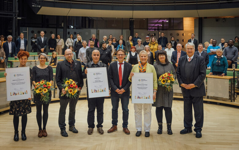 Sieben Personen auf einer Bühne halten Urkunden und Blumensträuße für den Sächsischen Integrationspreis, im Hintergrund weitere Gäste
